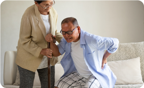 Photo of a senior woman sitting on a bench next to a walking trail in a park on a sunny day. She is holding her knee with a pained expression on her face.
