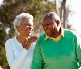 A senior couple are outside walking and smiling. The woman is looking at the man and putting her hand on his shoulder.